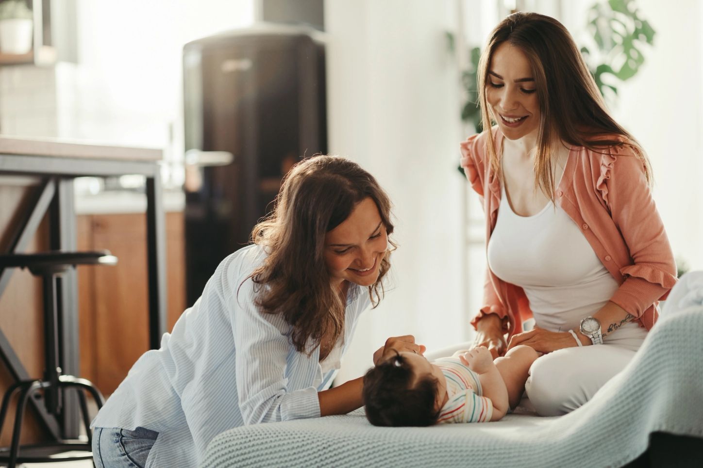 Two people with long brown hair gaze at a baby lying down on a cloth.