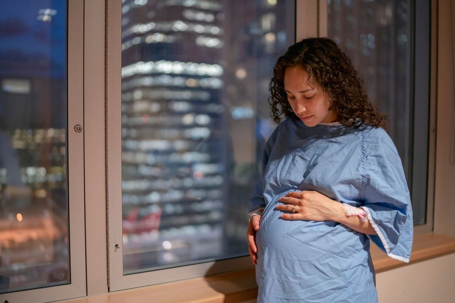 A person with long curly hair stares at their belly in a hospital window.