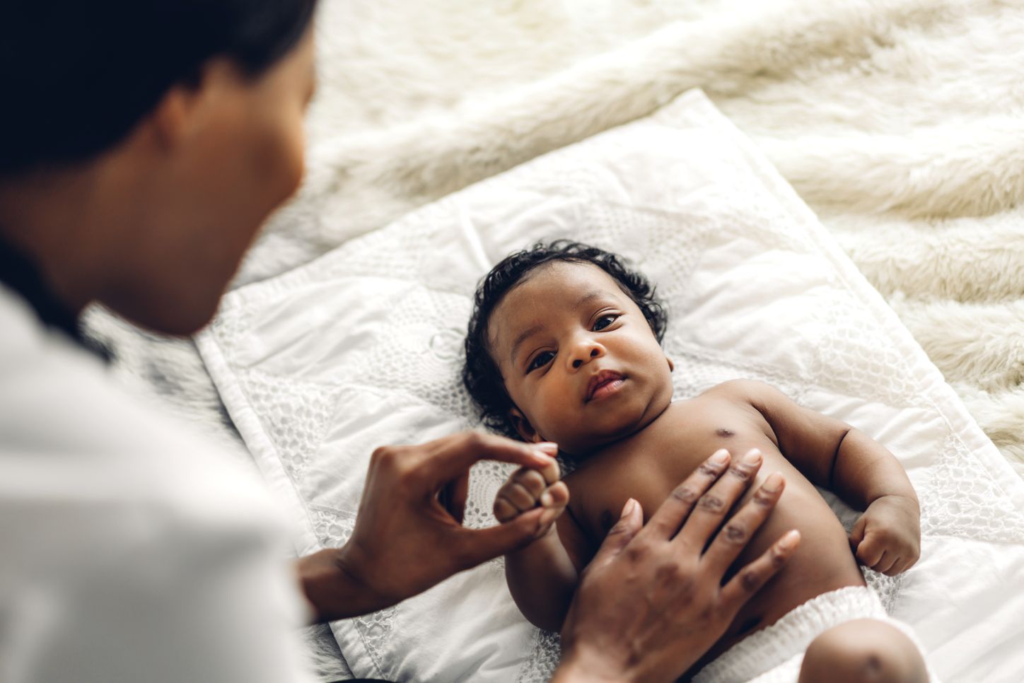 A naked baby with brown skin lies on a white towel. His fist is held by an adult hand and there is a hand on his stomach.