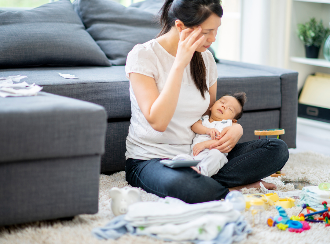 A tired-looking person sitting on the ground holds their sleeping baby.
