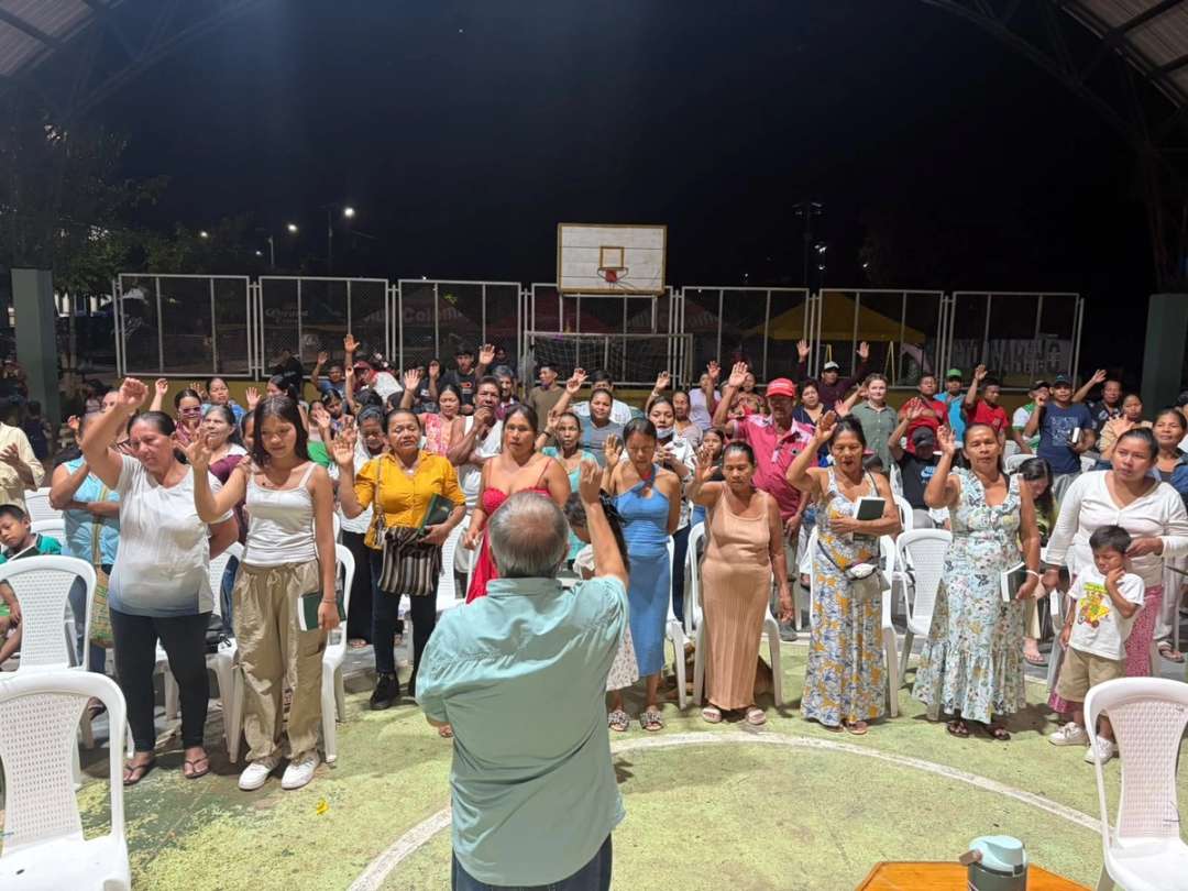 Mujeres, hombres y niños con las manos en alto durante una reunión nocturna en una cancha