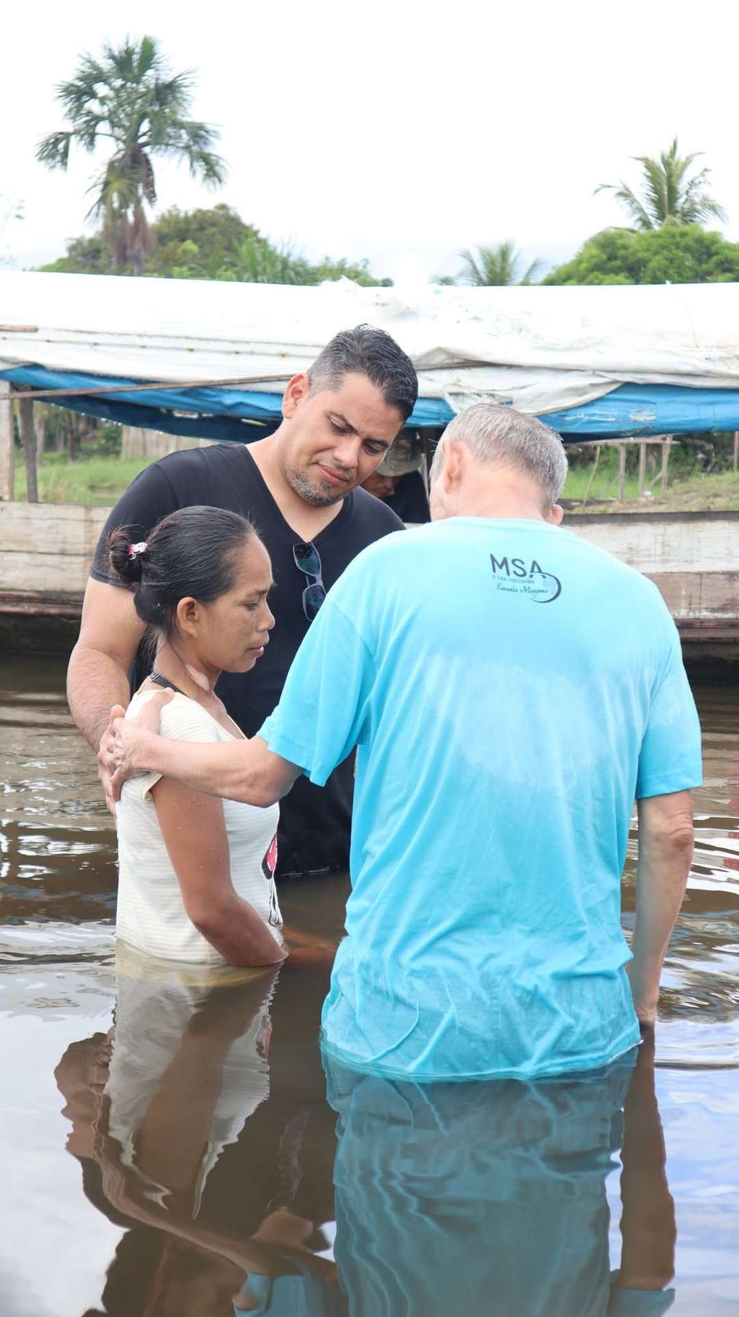 Un misionero bautiza a una joven en aguas del río junto a una comunidad palafítica
