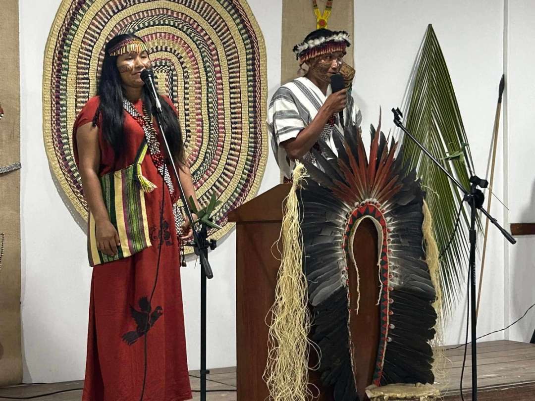 Una mujer y un hombre indígenas ante el micrófono, él con un gran tocado de plumas