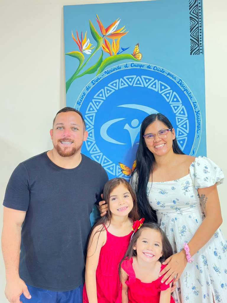 Edwin, Tamara y sus dos hijas sonríen frente a un mural con la declaración de propósito de JUCUM