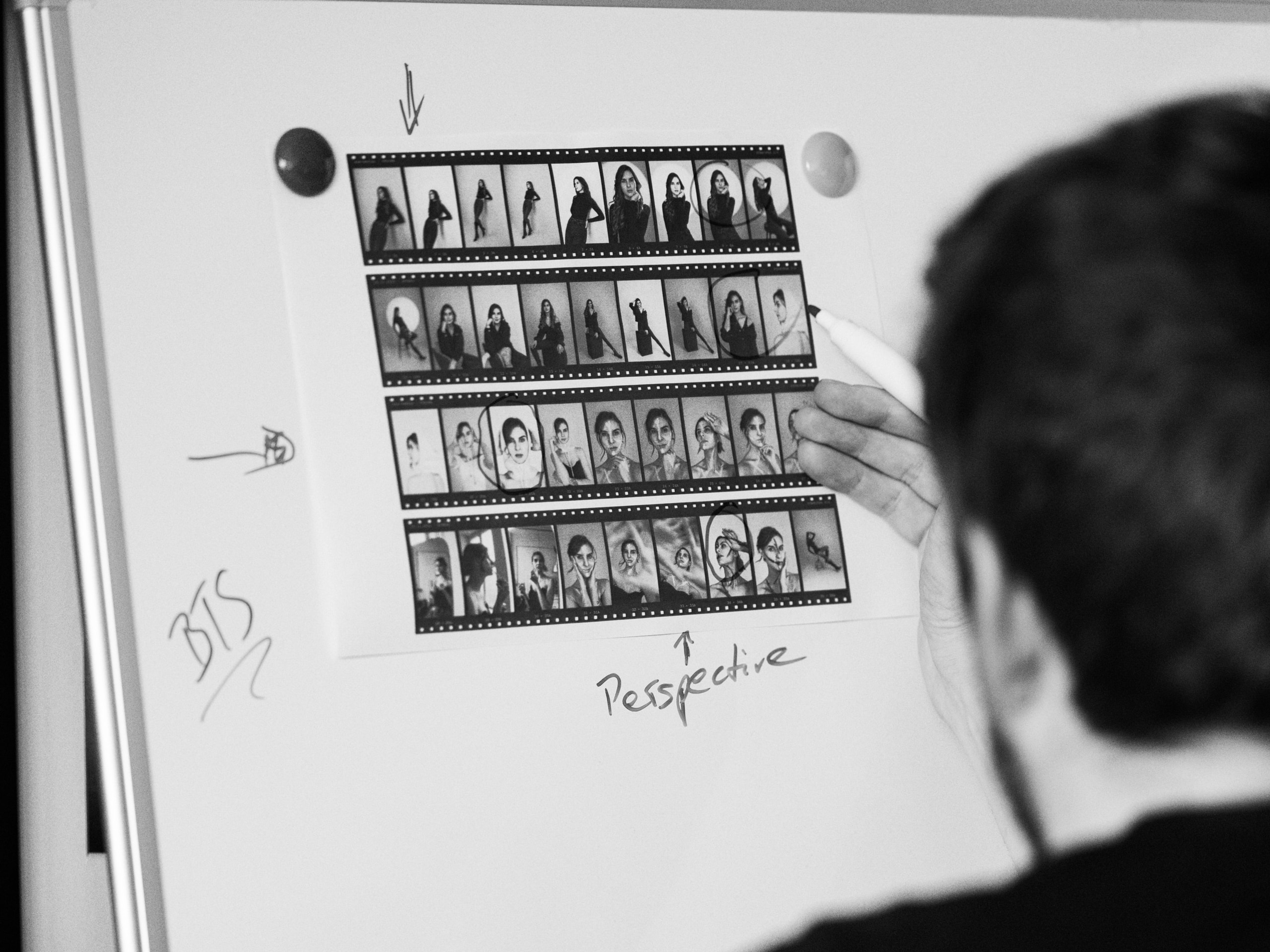 Schwarzweißer Kontaktbogen einer Portraitserie an einem Whiteboard, mehrere Bilder mit Stift eingekreist, Notizen „BTS" und „Perspective" daneben. Eine Hand mit Stift markiert gerade ein Bild, im Vordergrund unscharf der Hinterkopf einer Person.