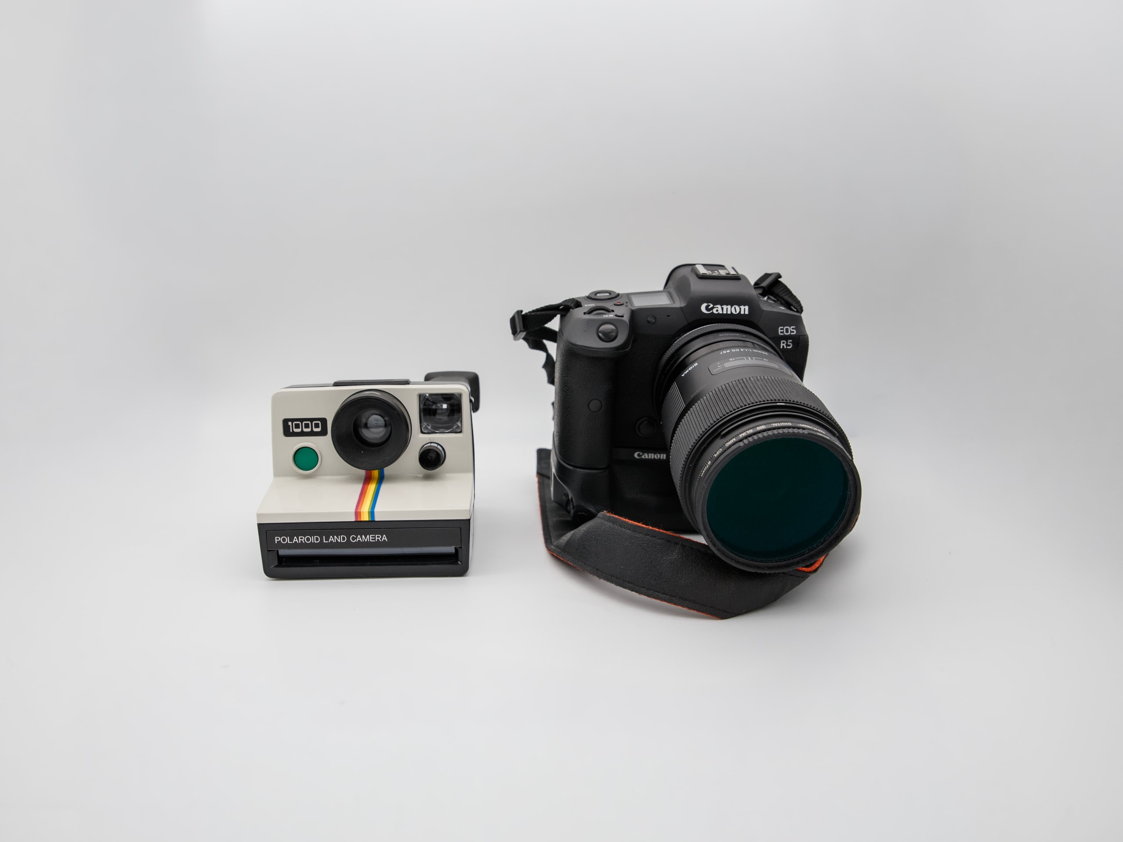 Polaroid Land Camera 1000 mit Regenbogenstreifen neben einer Canon EOS R5 mit großem Objektiv auf hellgrauem Hintergrund. Zwei Kameras mit derselben Funktion und gegensätzlicher Form.