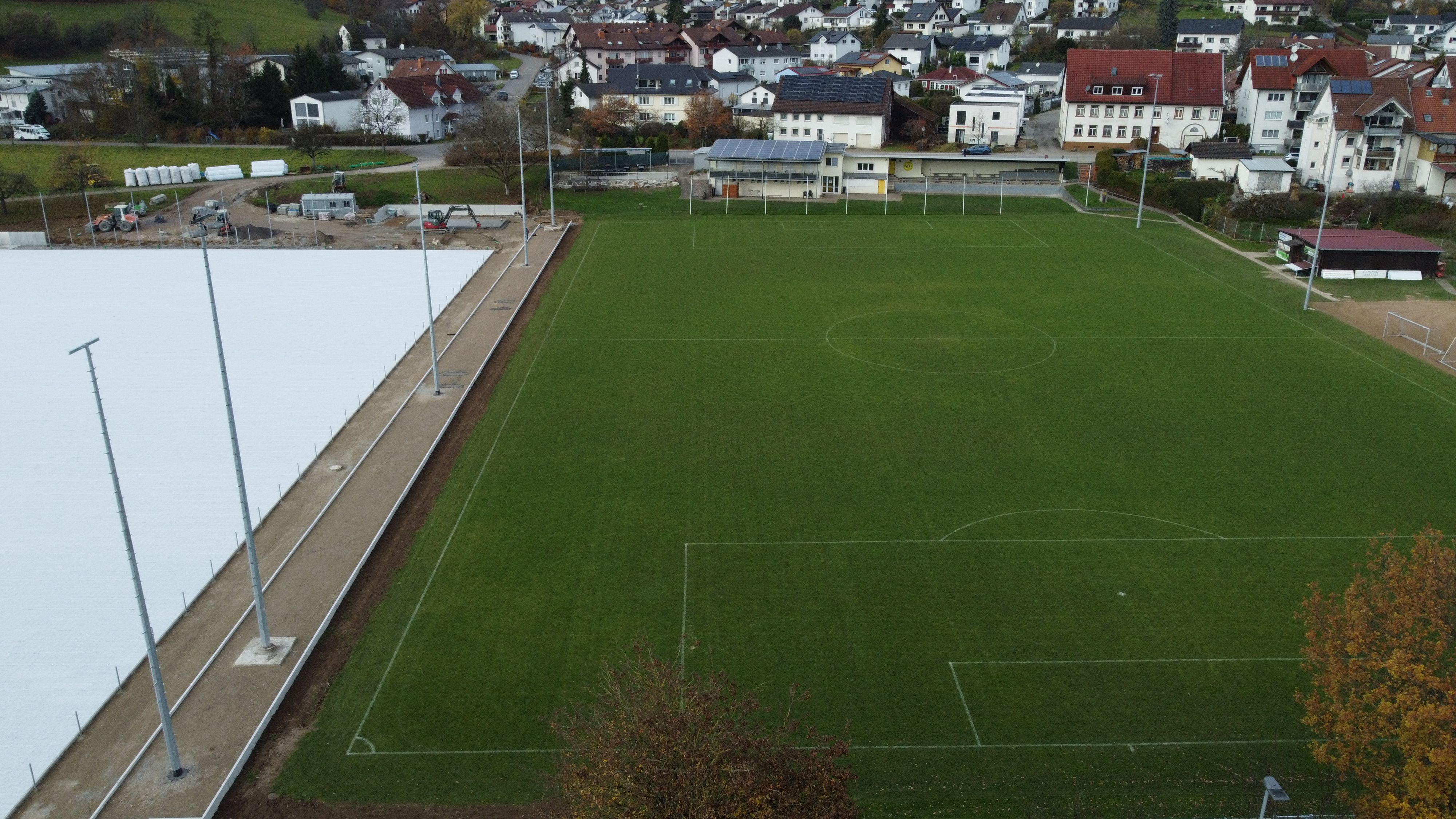 Luftaufnahme mit dem neuen Kunstrasenplatz links und dem bestehenden Rasenplatz mit Vereinsheim rechts nebeneinander