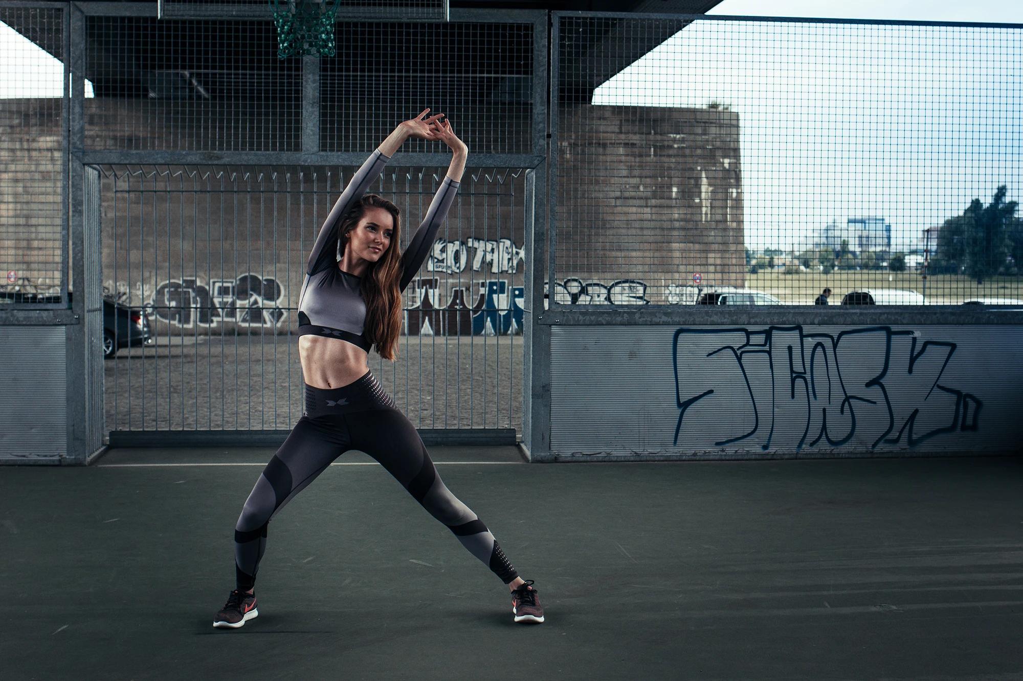 Brooke beim Stretching auf einem Bolzplatz mit Gitterzaun und Graffiti in Düsseldorf