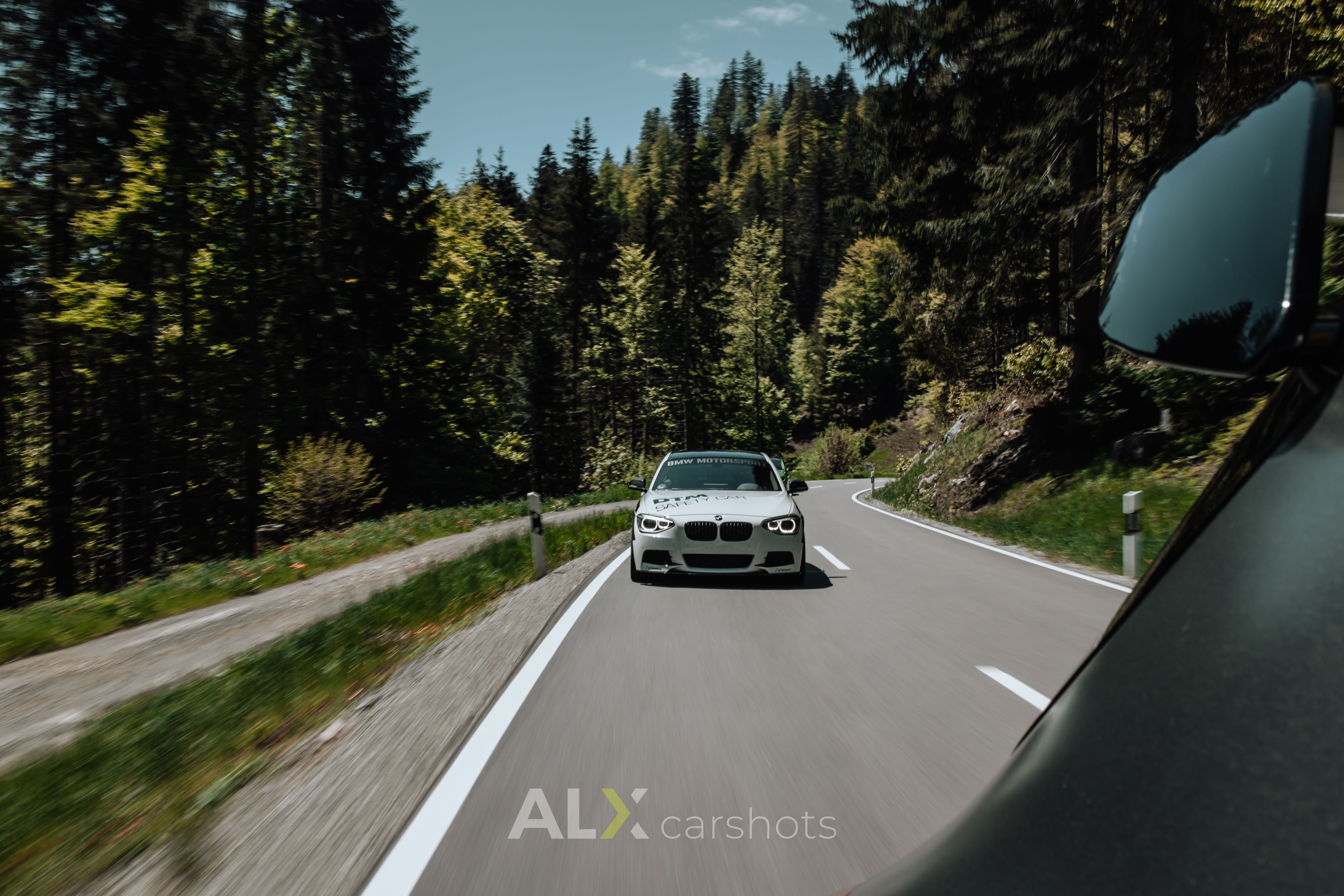 Weißer BMW 1er als Rollingshot, aufgenommen aus einem Begleitfahrzeug mit Außenspiegel im Vordergrund
