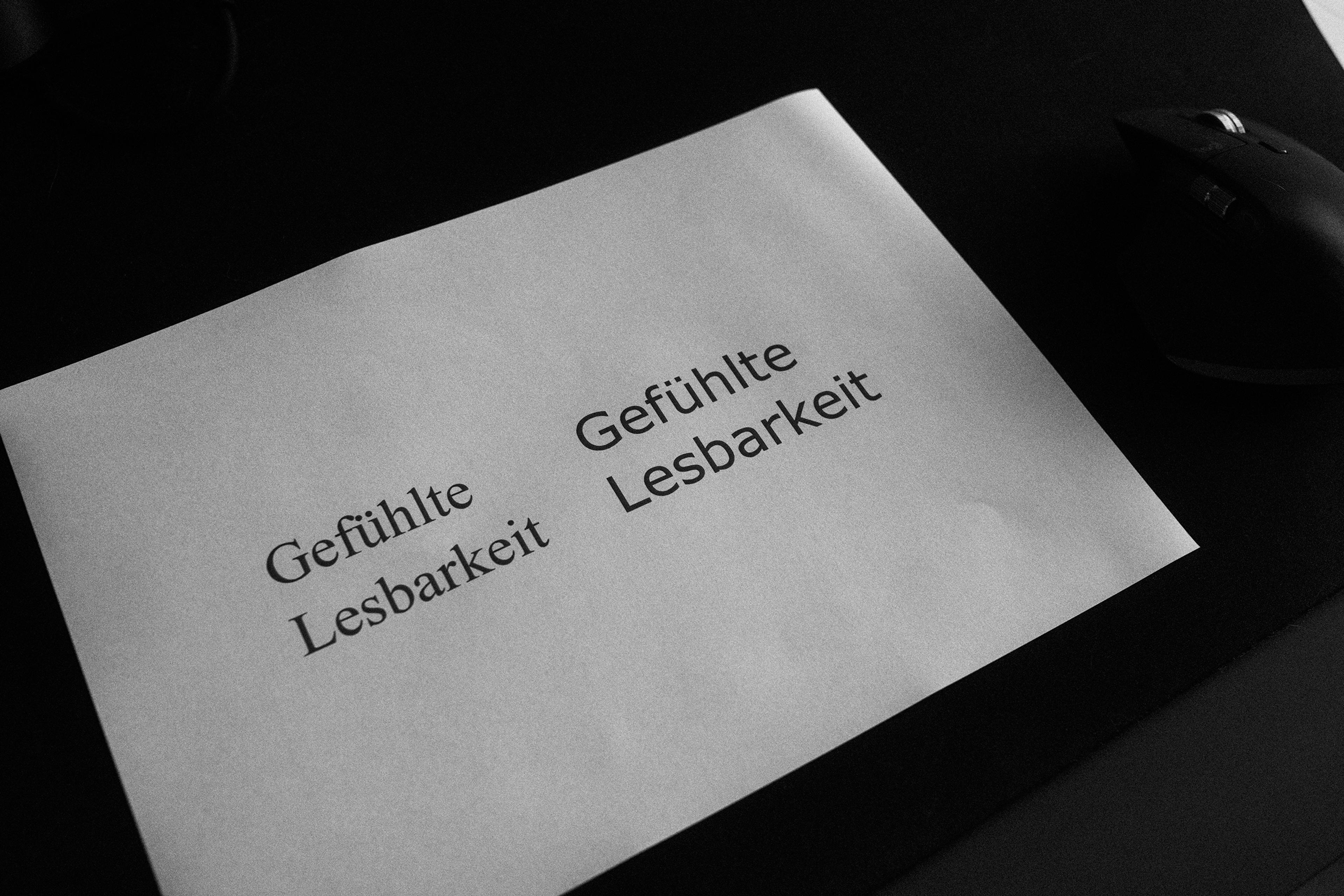 Blatt Papier mit den Worten „Gefühlte Lesbarkeit", links in einer Serifenschrift, rechts in einer serifenlosen Schrift, auf dunklem Schreibtisch.