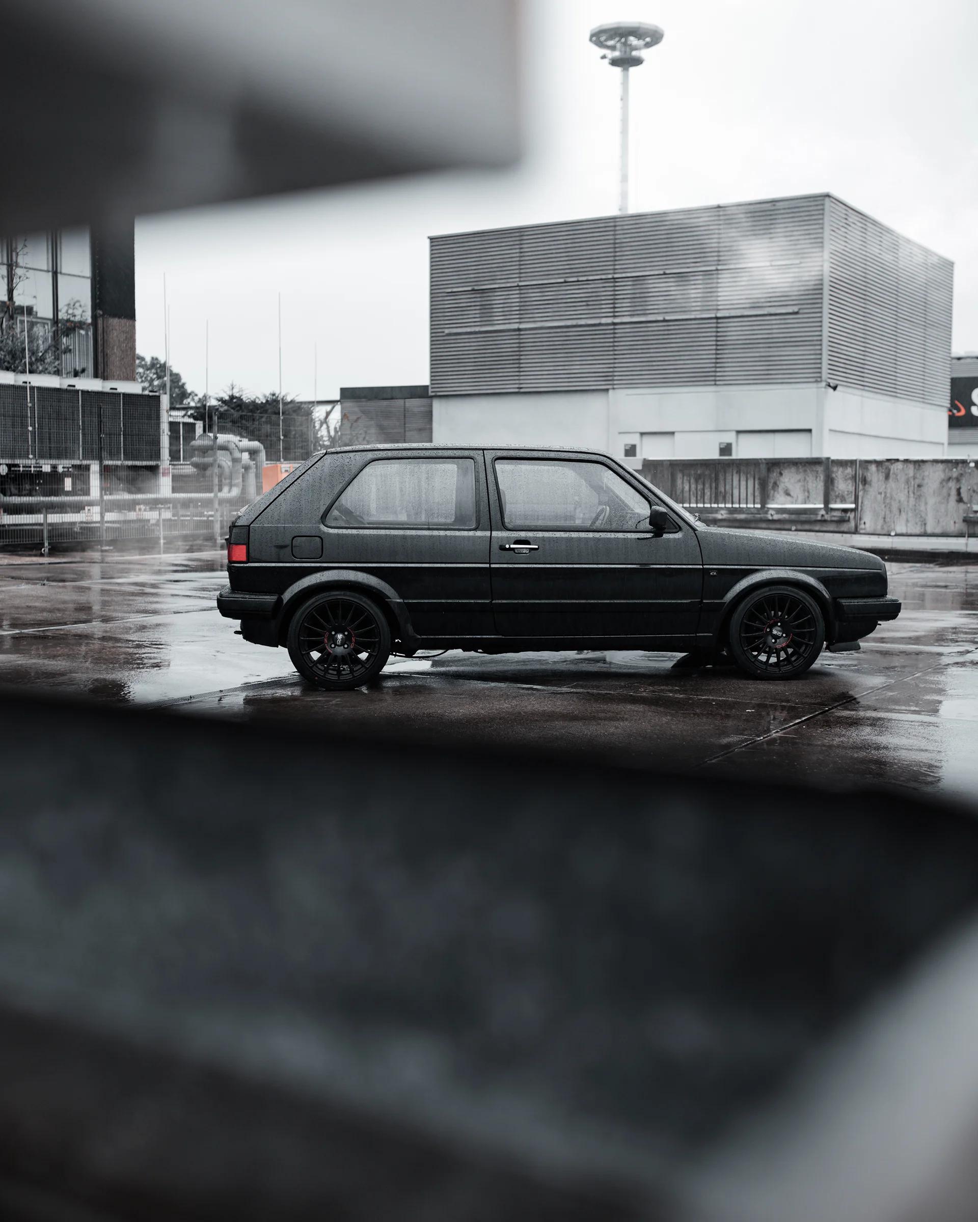 Tiefergelegter schwarzer VW Golf II im Seitenprofil auf regennassem Asphalt vor einer Industriekulisse.