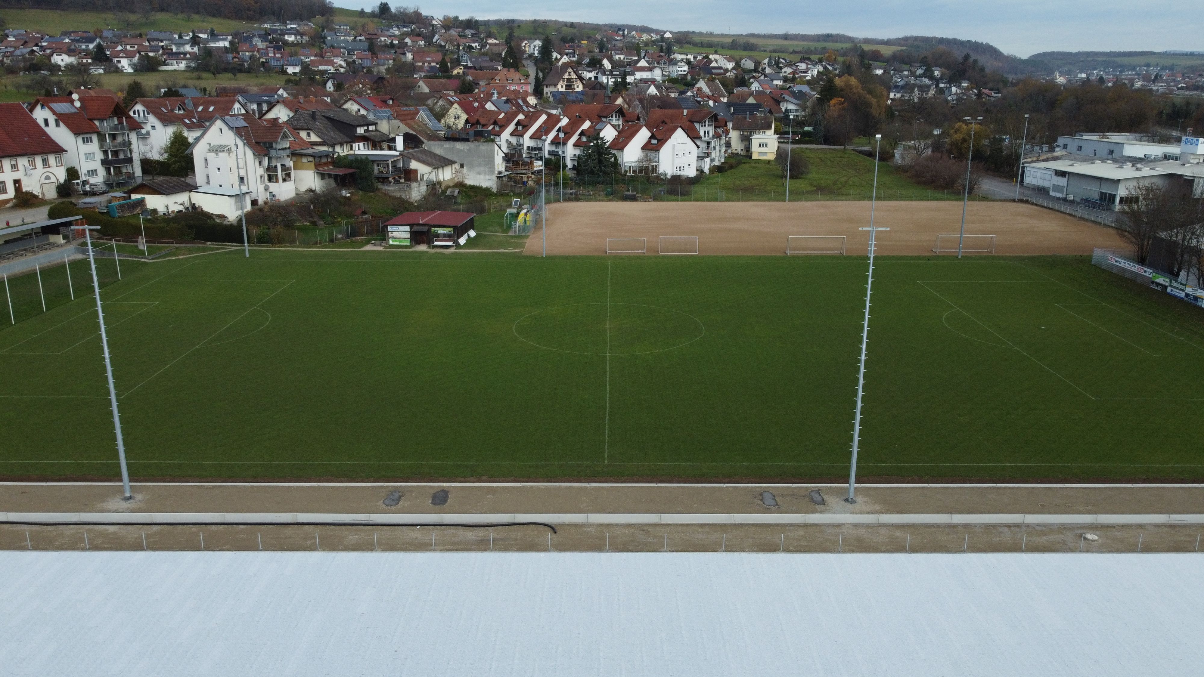 Blick über den bestehenden grünen Rasenplatz auf die weiße Baufläche des neuen Kunstrasens und den alten Hartplatz dahinter