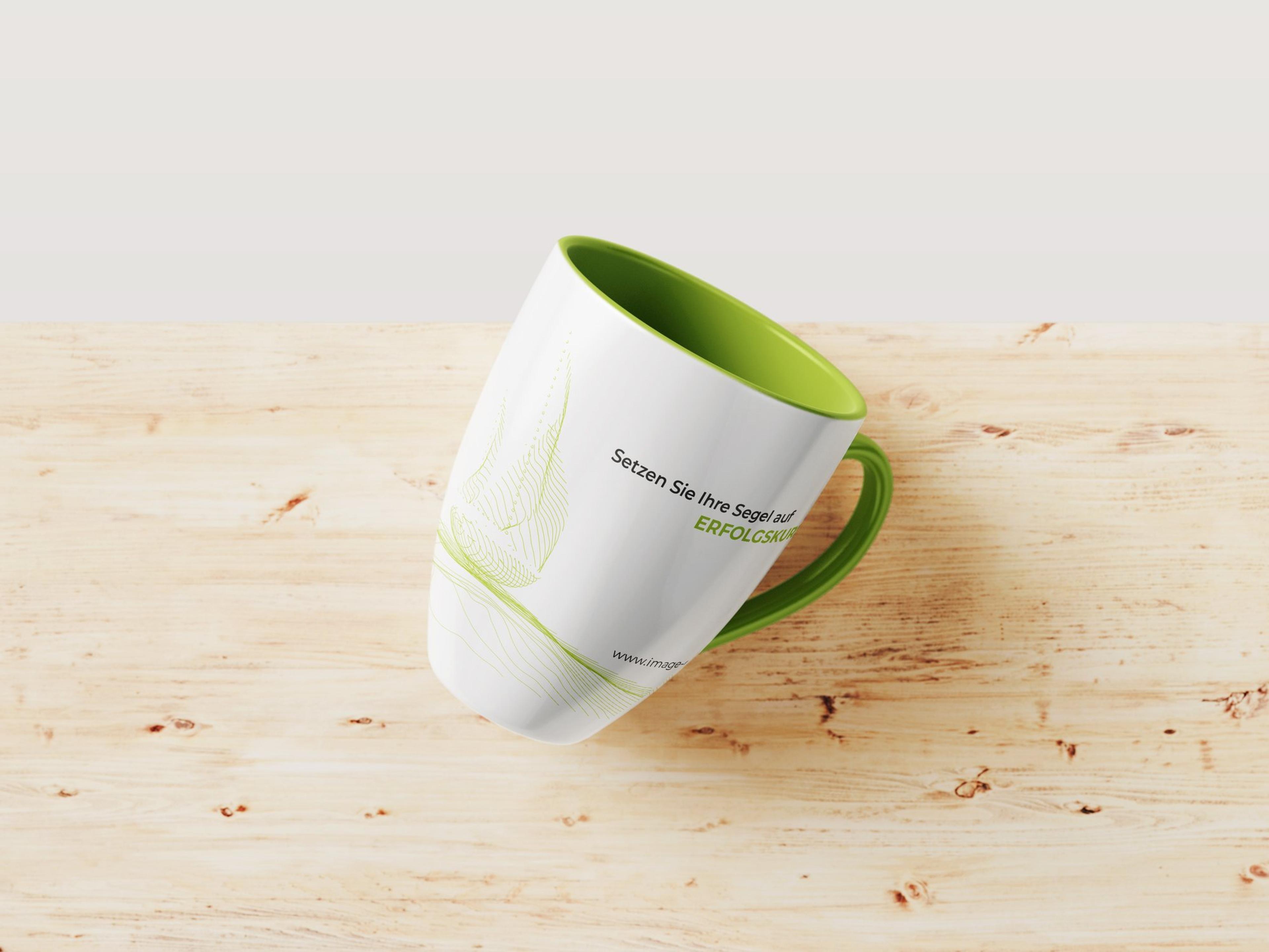 Weiße Werbetasse von Image Digital mit grünem Innenraum und Henkel auf einem Holztisch, mit dem Spruch Setzen Sie Ihre Segel auf Erfolgskurs.