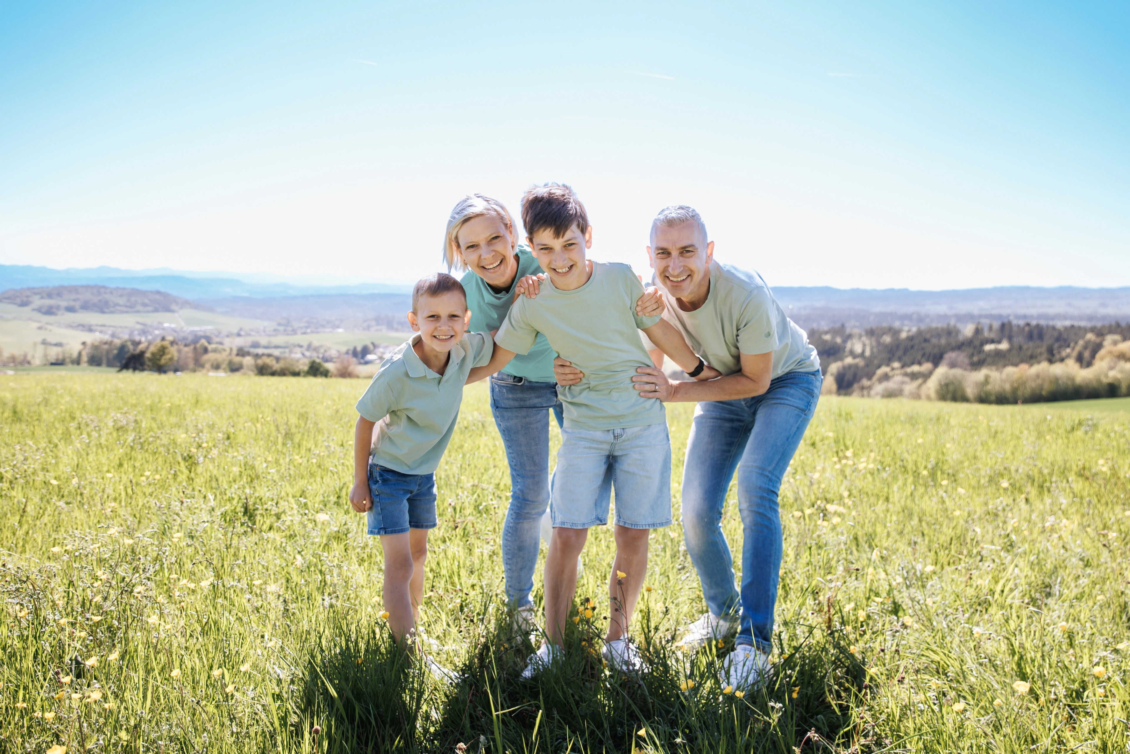 Familie mit zwei Kindern in enger Umarmung auf einer Frühlingswiese über dem Hochrhein mit weitem Blick über die Hügel, Familienfotografie von aLx-design