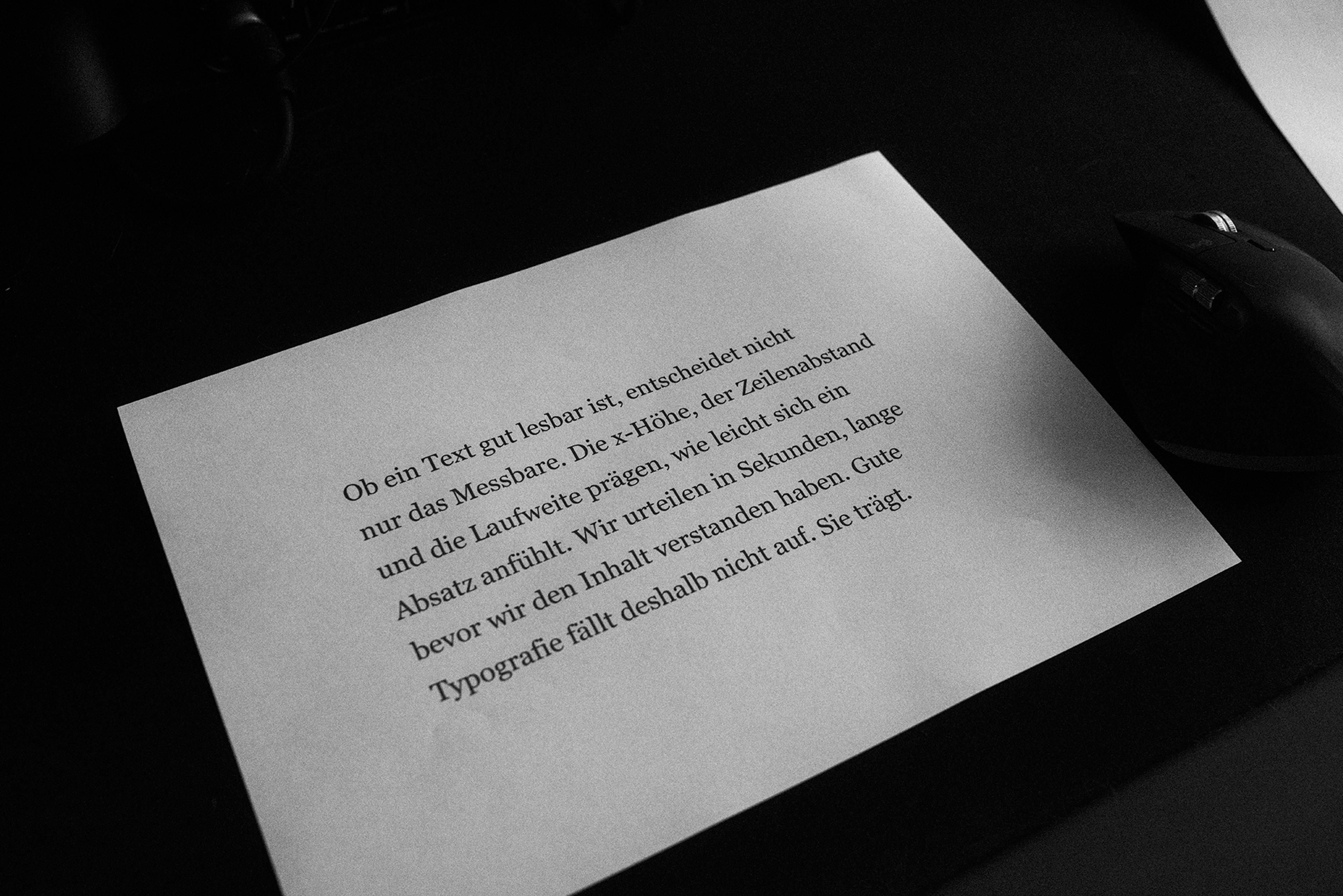 Gedruckter Fließtextabsatz in einer Serifenschrift auf hellem Papier, daneben eine Computermaus auf dunklem Schreibtisch.