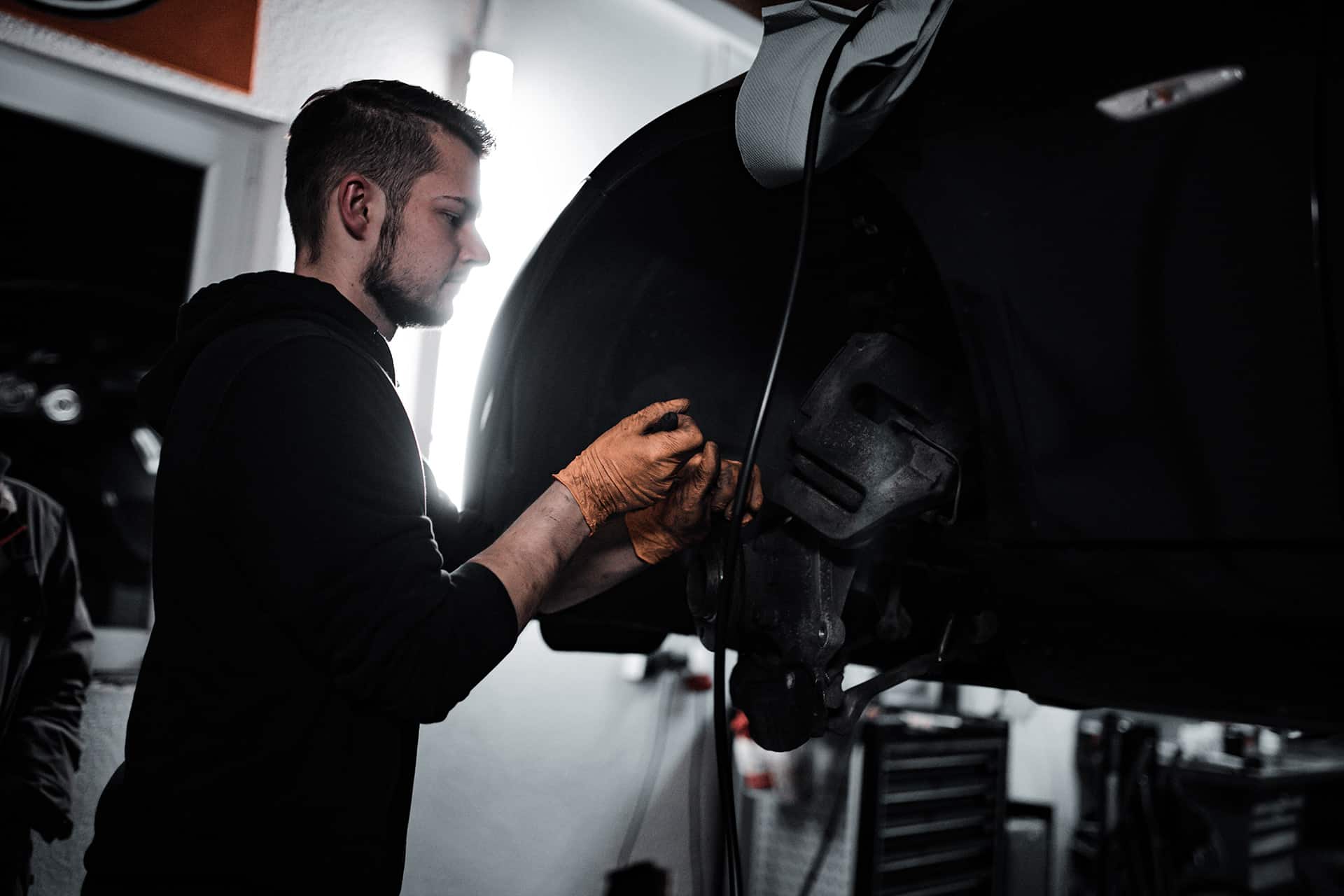 Kfz-Mechaniker in dunklem Hoodie arbeitet an der freigelegten Vorderradbremse eines aufgebockten Fahrzeugs, Werkstatt im Streiflicht.
