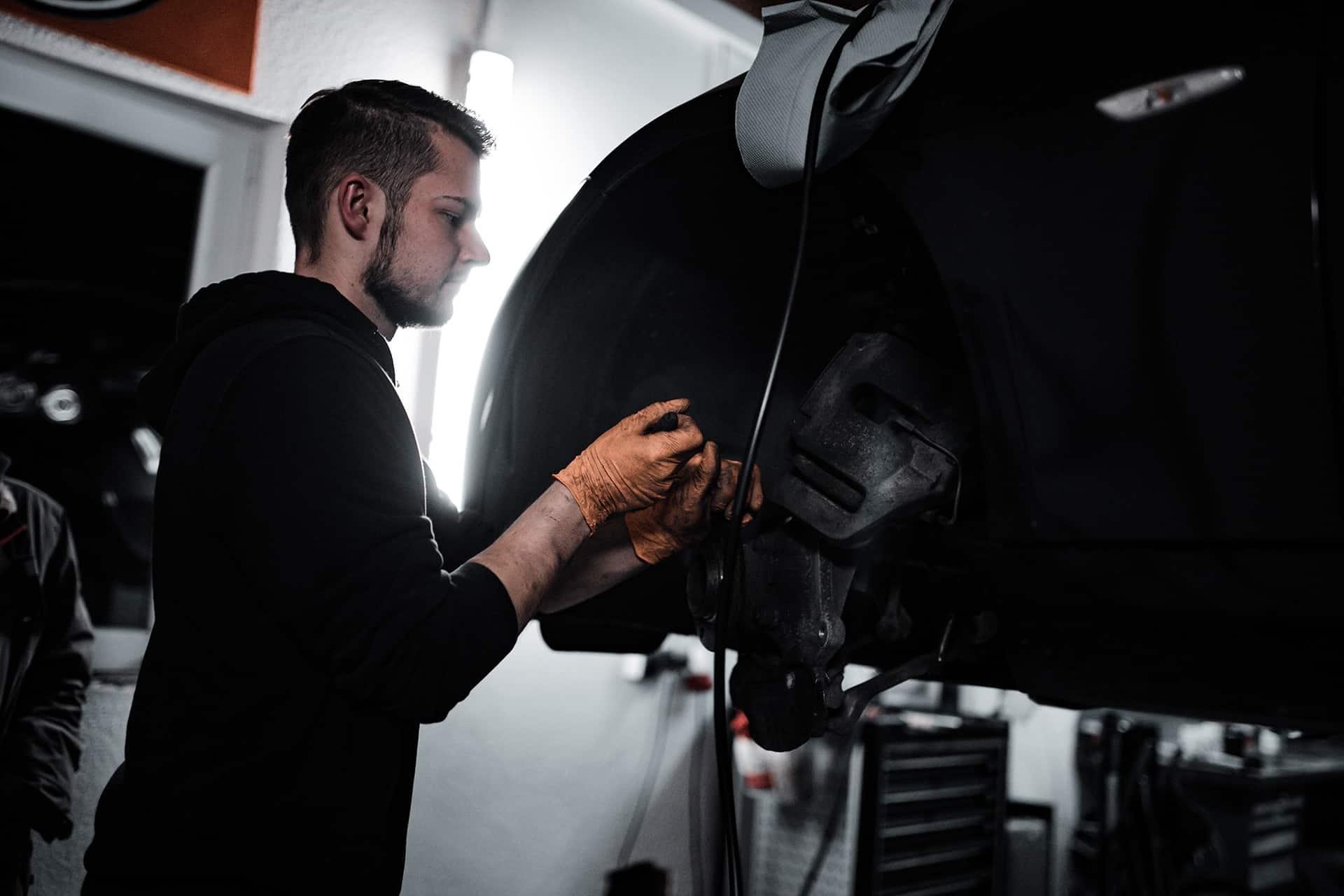 Kfz-Mechaniker in dunklem Hoodie arbeitet an der freigelegten Vorderradbremse eines aufgebockten Fahrzeugs, Werkstatt im Streiflicht.
