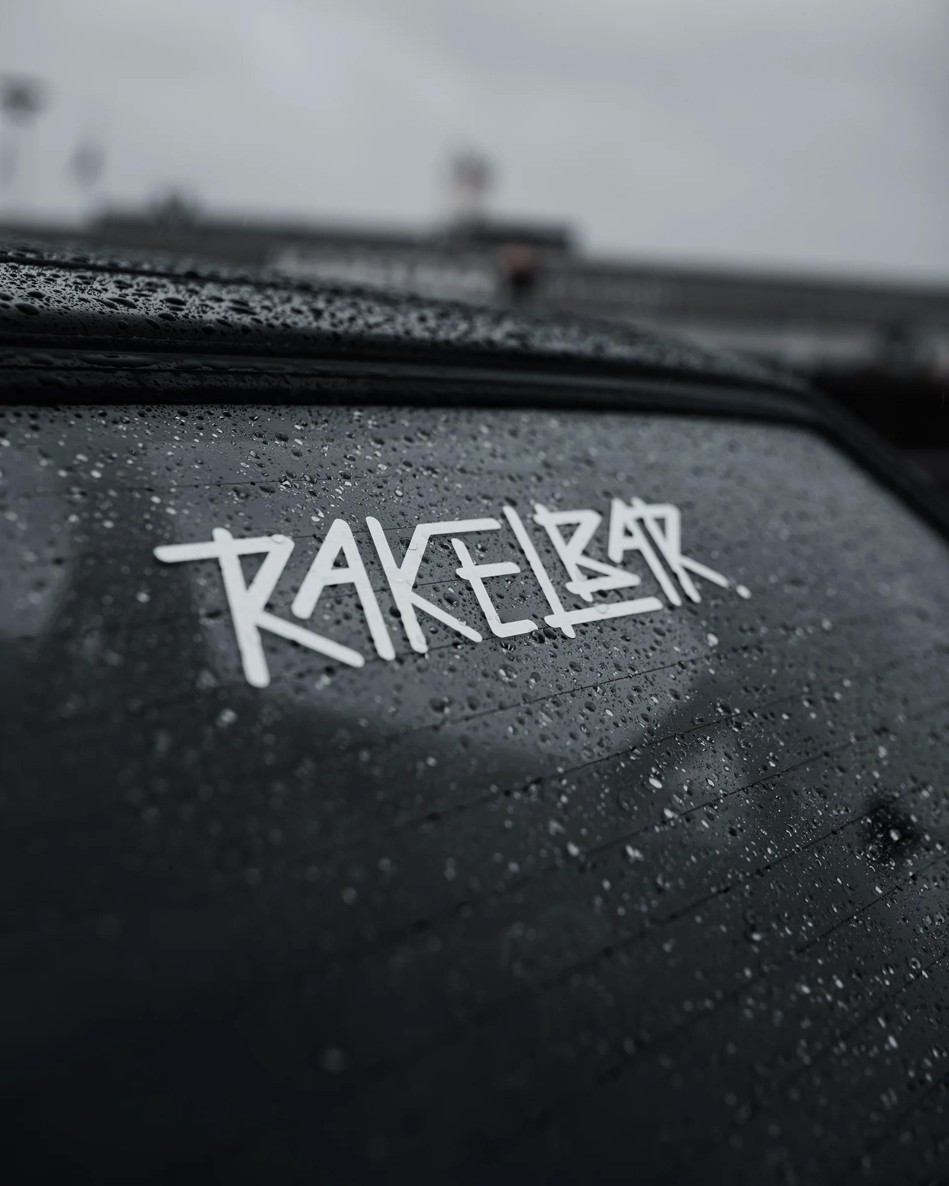 Der weiße Rakelbar-Schriftzug als Aufkleber auf einer regennassen Fahrzeugscheibe.