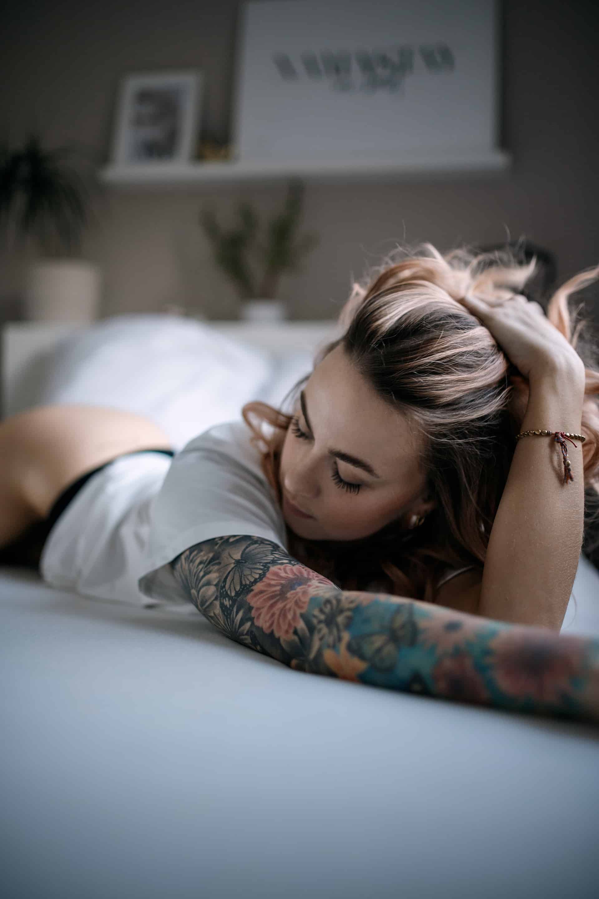 Zintle im weißen Shirt bäuchlings auf dem Bett, Hände im Haar, Tattoos am Arm