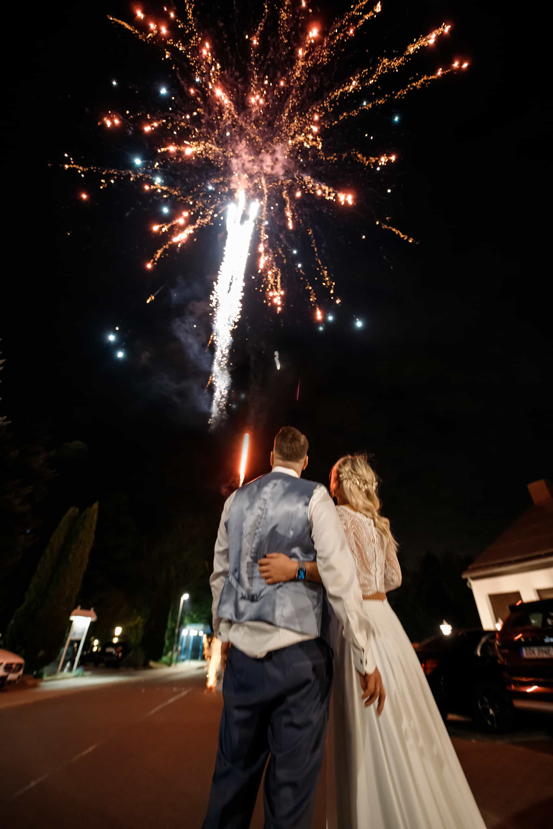 Brautpaar von hinten Arm in Arm beim Feuerwerk am Abend, goldene Funken über dem dunklen Himmel vor der Location.