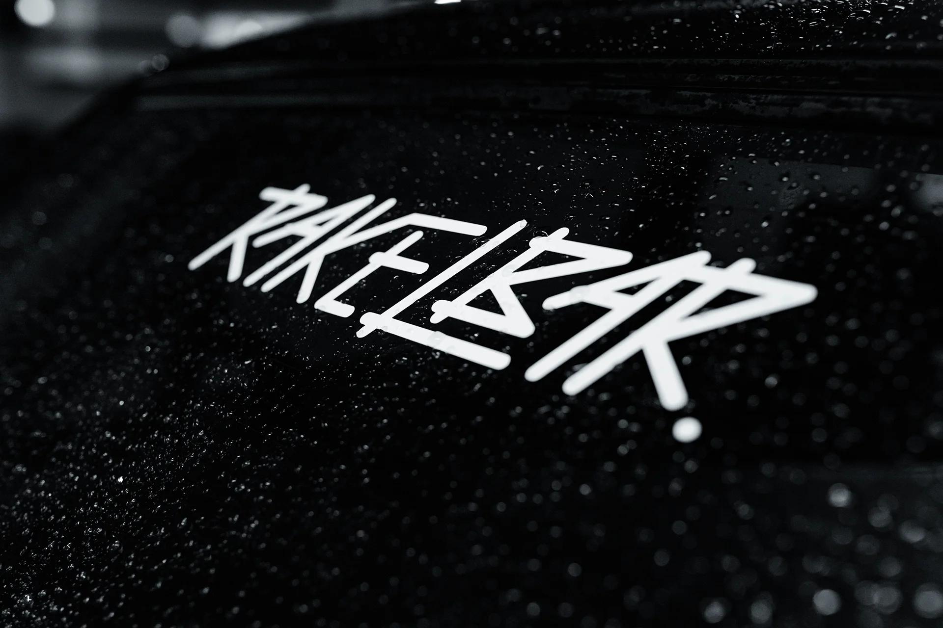Detailaufnahme des Rakelbar-Aufklebers auf der regennassen schwarzen Lackfläche eines Autos.