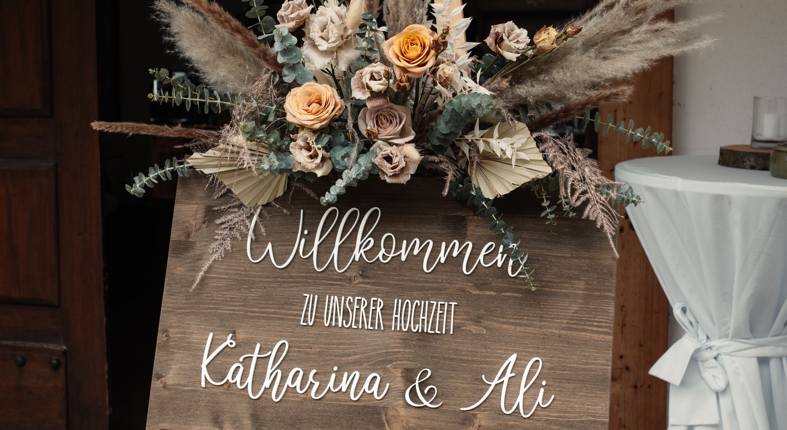 Hölzernes Willkommensschild mit Trockenblumen und dem Schriftzug Katharina und Ali