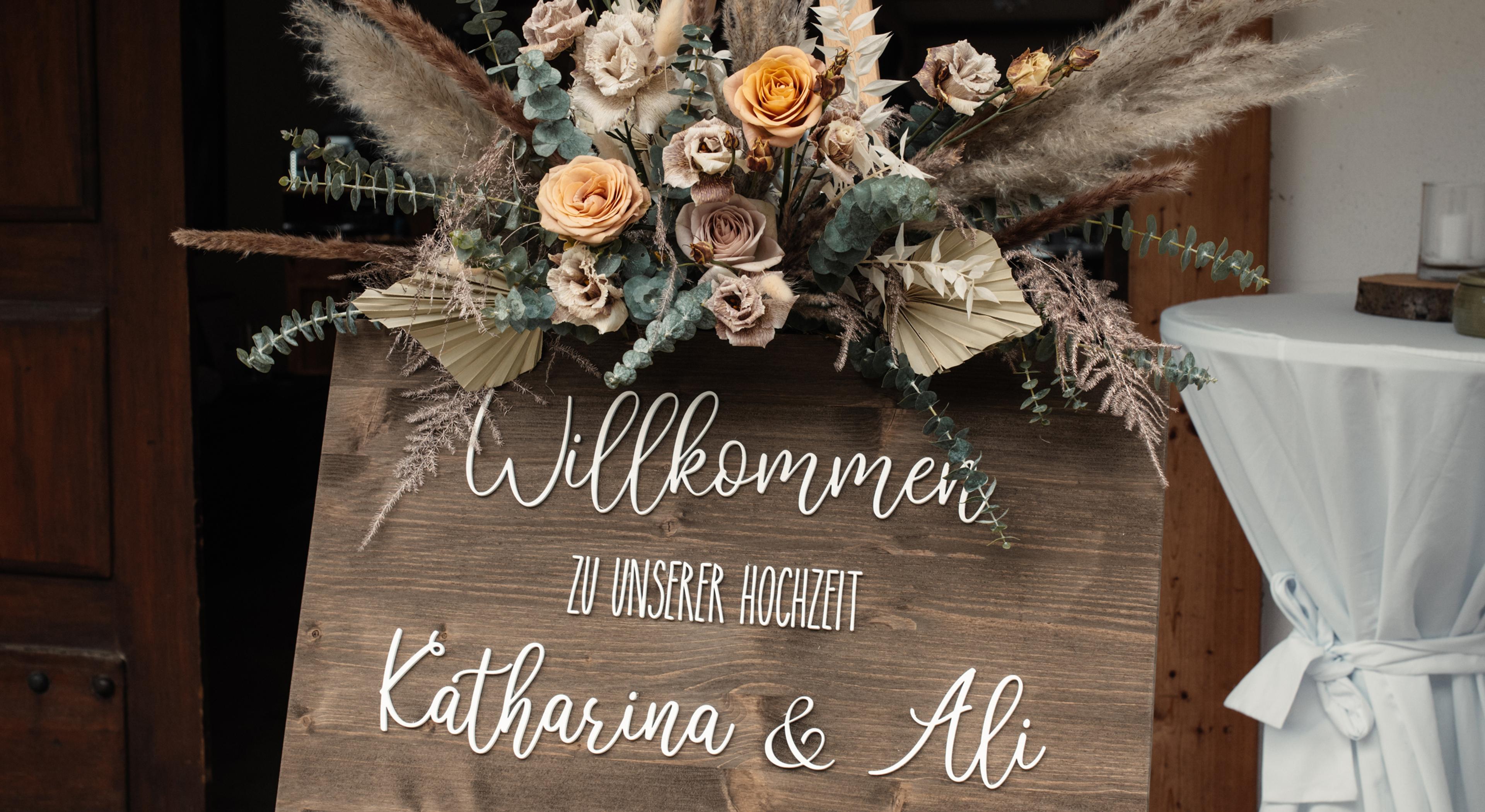 Hölzernes Willkommensschild mit Trockenblumen und dem Schriftzug Katharina und Ali