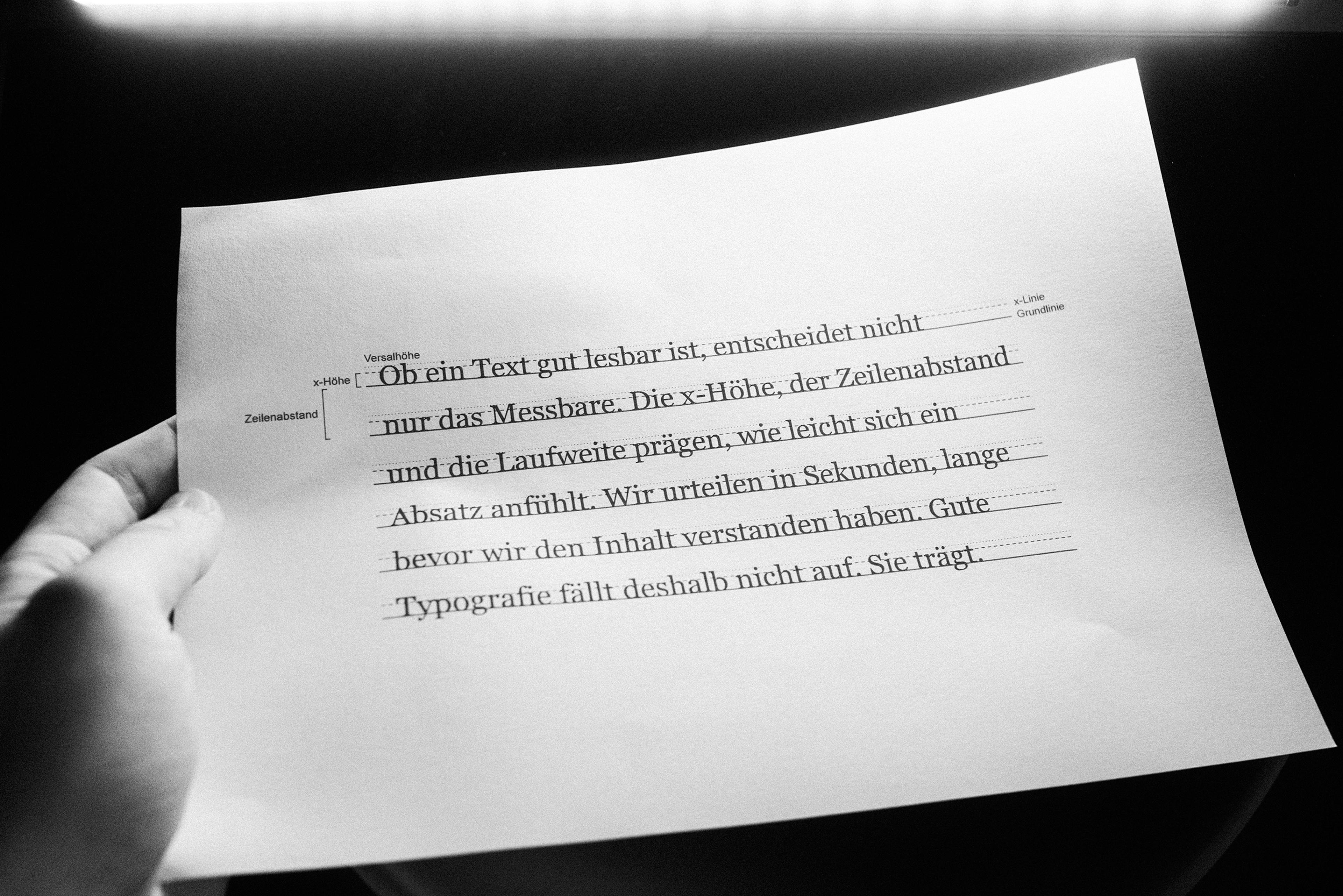 Hand hält ein Blatt mit gedrucktem Textabsatz, darauf eingezeichnete Hilfslinien für Versalhöhe, x-Höhe, Grundlinie und Zeilenabstand.