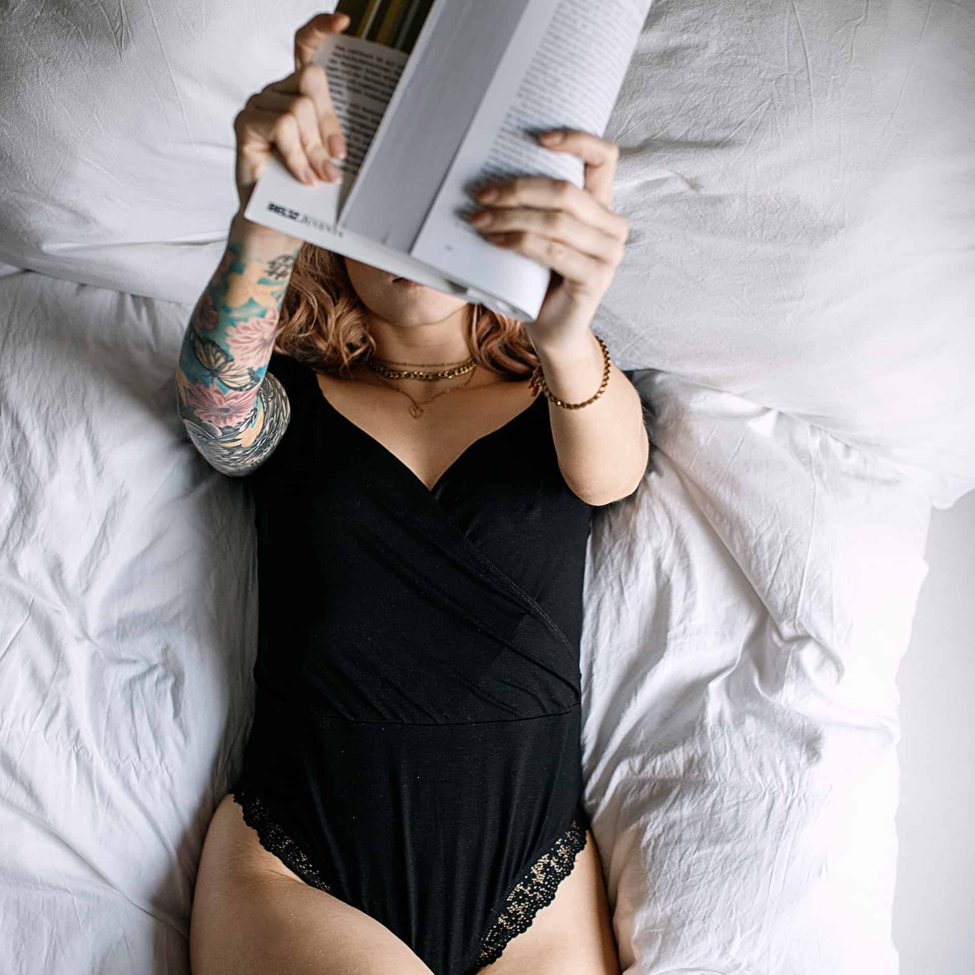 Zintle im schwarzen Body auf weißer Bettwäsche, ein aufgeschlagenes Buch vor dem Gesicht