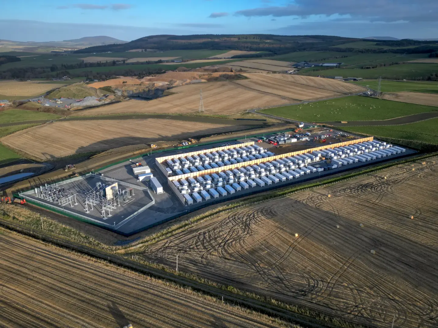 Aerial view of a large-scale battery energy storage facility with rows of white containers and an electrical substation, surrounded by agricultural fields.