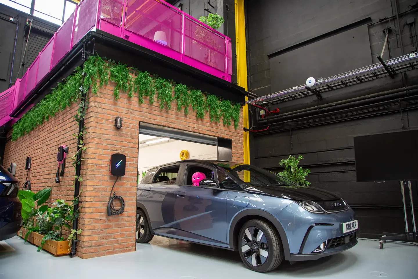 A blue-gray electric car with a pink figure inside partially exits a brick garage with an EV charger, beneath a vibrant pink balcony.