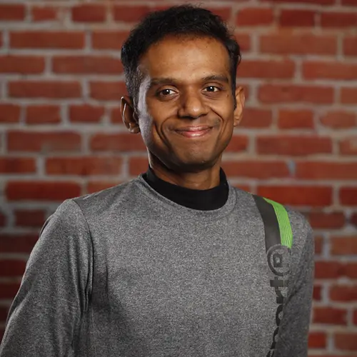 A smiling man with dark hair in a grey long-sleeve shirt with a green stripe on the shoulder, against a brick wall background.