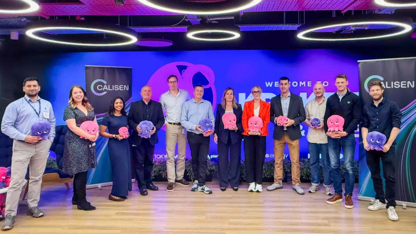 Twelve people stand in a row in front of a Calisen banner, each holding a pink or purple plush octopus toy.