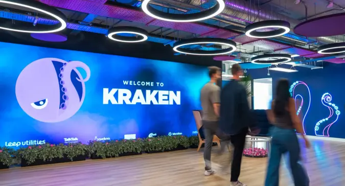 A modern office reception with a large screen displaying "WELCOME TO KRAKEN" and a purple octopus logo, with blurred people walking.