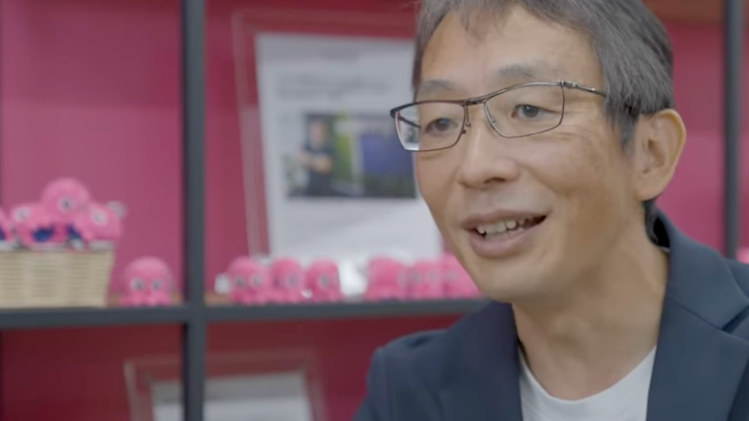 A man with glasses smiles in front of a pink shelf displaying small pink octopus toys.