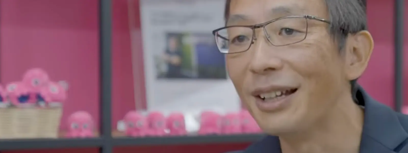 A man with glasses smiles in front of a pink shelf displaying small pink octopus toys.