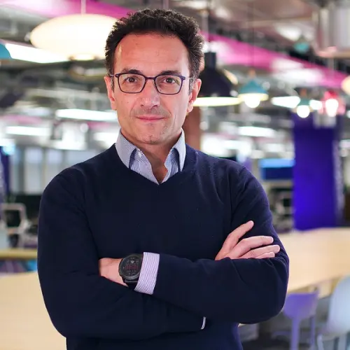 A man in glasses and a blue sweater with crossed arms, wearing a smartwatch, stands in a vibrant office.