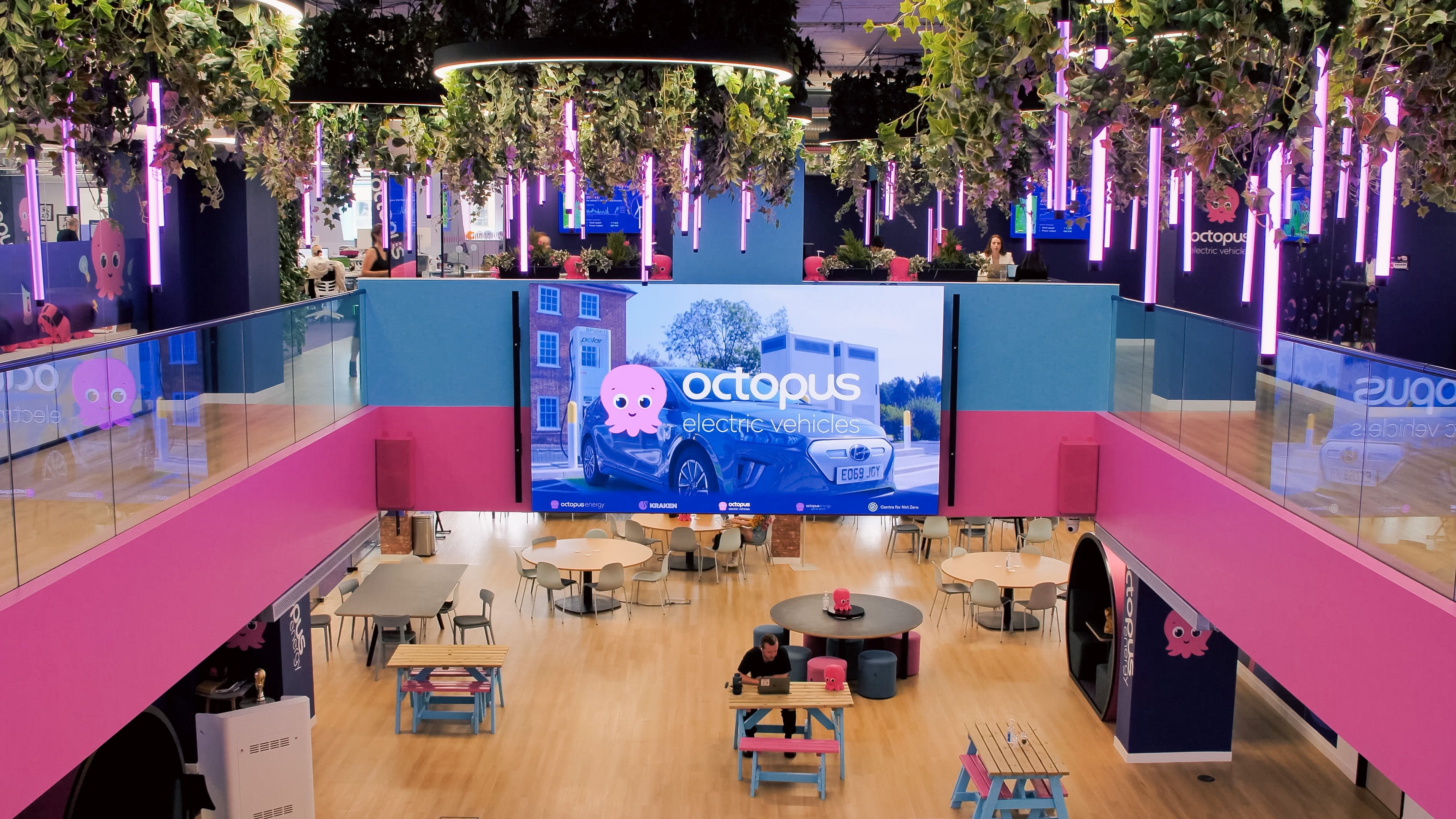 A vibrant, multi-level office with pink and blue decor, hanging plants, purple neon lights, and a large screen advertising Octopus Electric Vehicles.