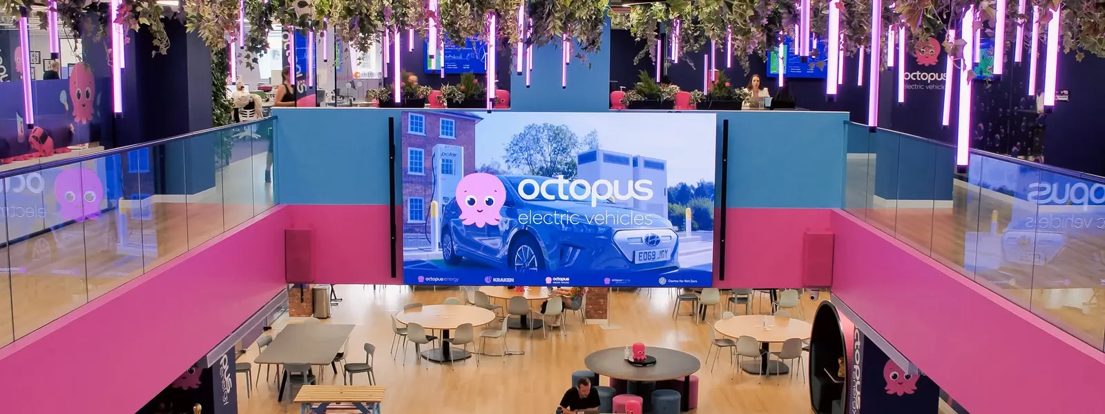 A vibrant, multi-level office with pink and blue decor, hanging plants, purple neon lights, and a large screen advertising Octopus Electric Vehicles.