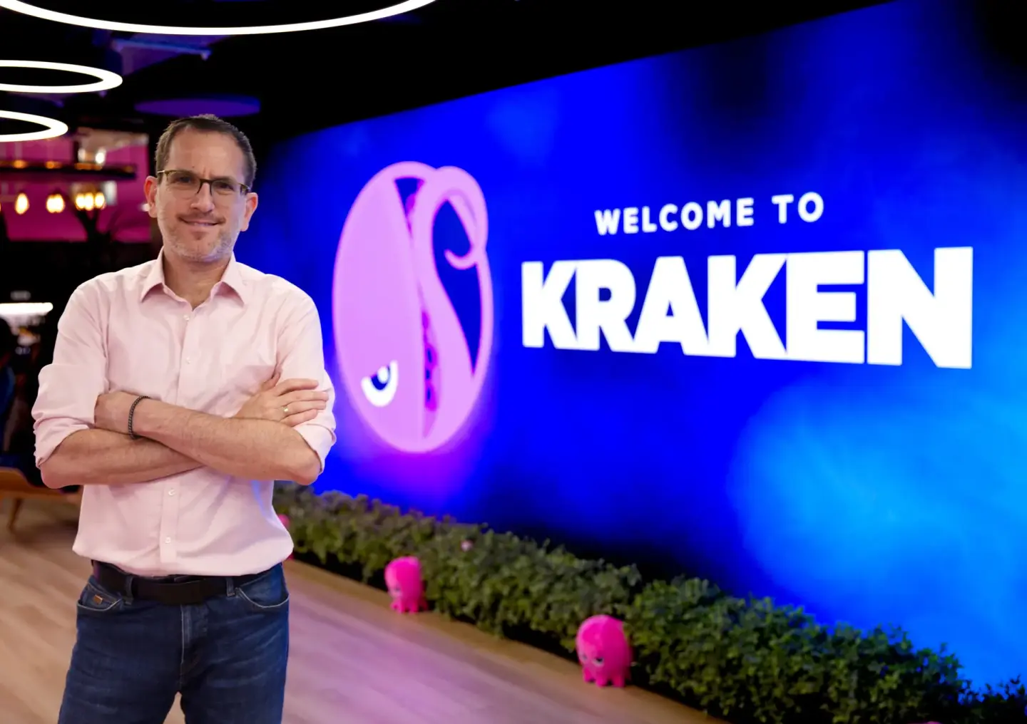 A man in glasses and a pink shirt smiles with crossed arms in front of a blue wall featuring the Kraken logo and "Welcome to Kraken" text.