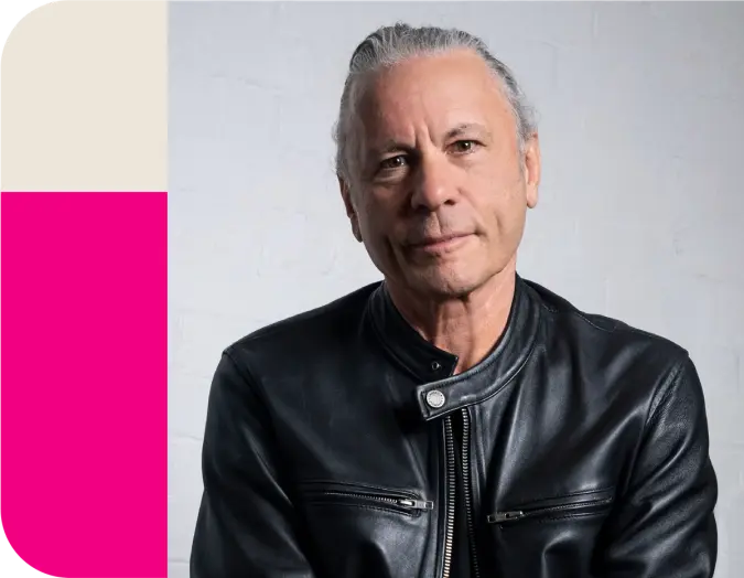Bruce Dickinson, with slicked-back grey hair, wearing a black leather jacket, smiles.