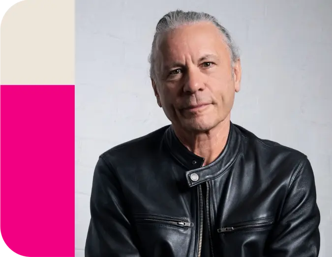 Bruce Dickinson, with slicked-back grey hair, wearing a black leather jacket, smiles.