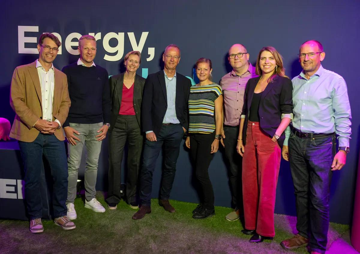 Eight professionals, four men and four women, stand on artificial grass in front of a blue wall with "Energy," text.