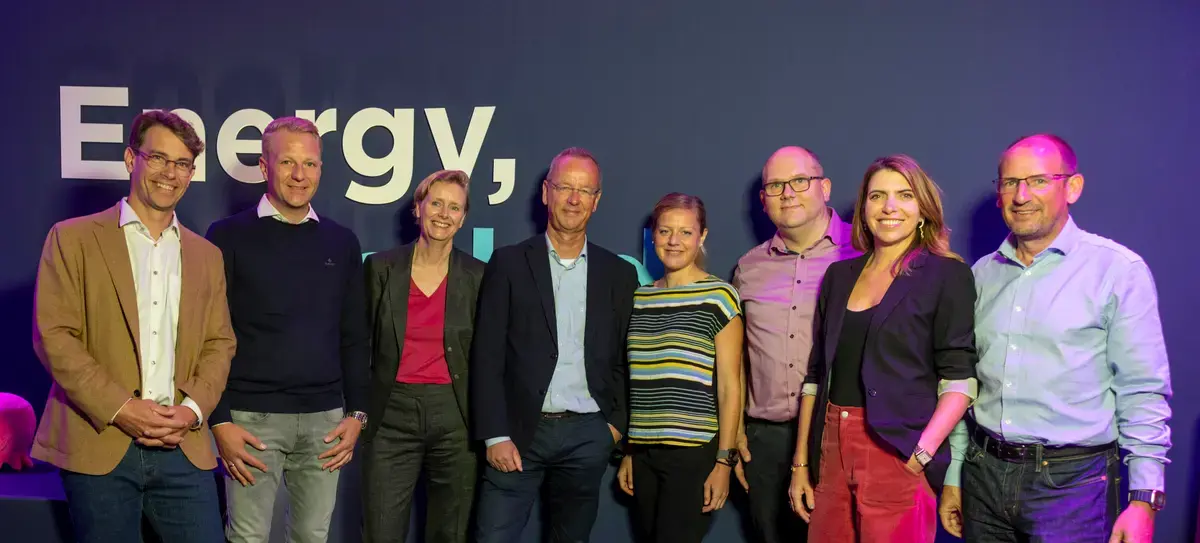Eight professionals, four men and four women, stand on artificial grass in front of a blue wall with "Energy," text.