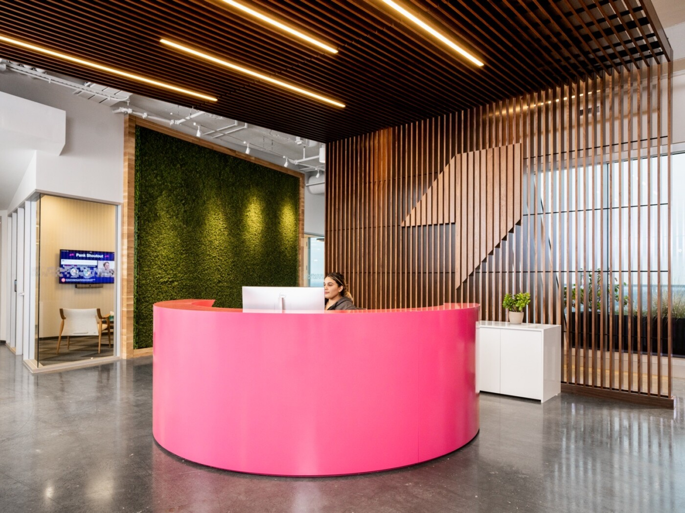 A modern reception area with a bright pink curved desk, a woman working, a large moss wall, and wood slat walls and ceiling.