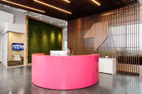 A modern reception area with a bright pink curved desk, a woman working, a large moss wall, and wood slat walls and ceiling.