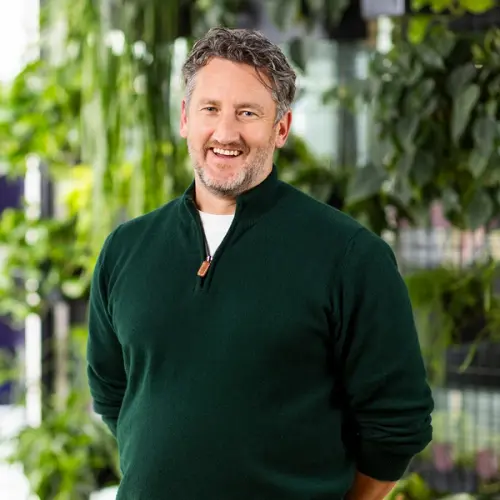 A smiling man in a green sweater stands in front of a green plant wall.