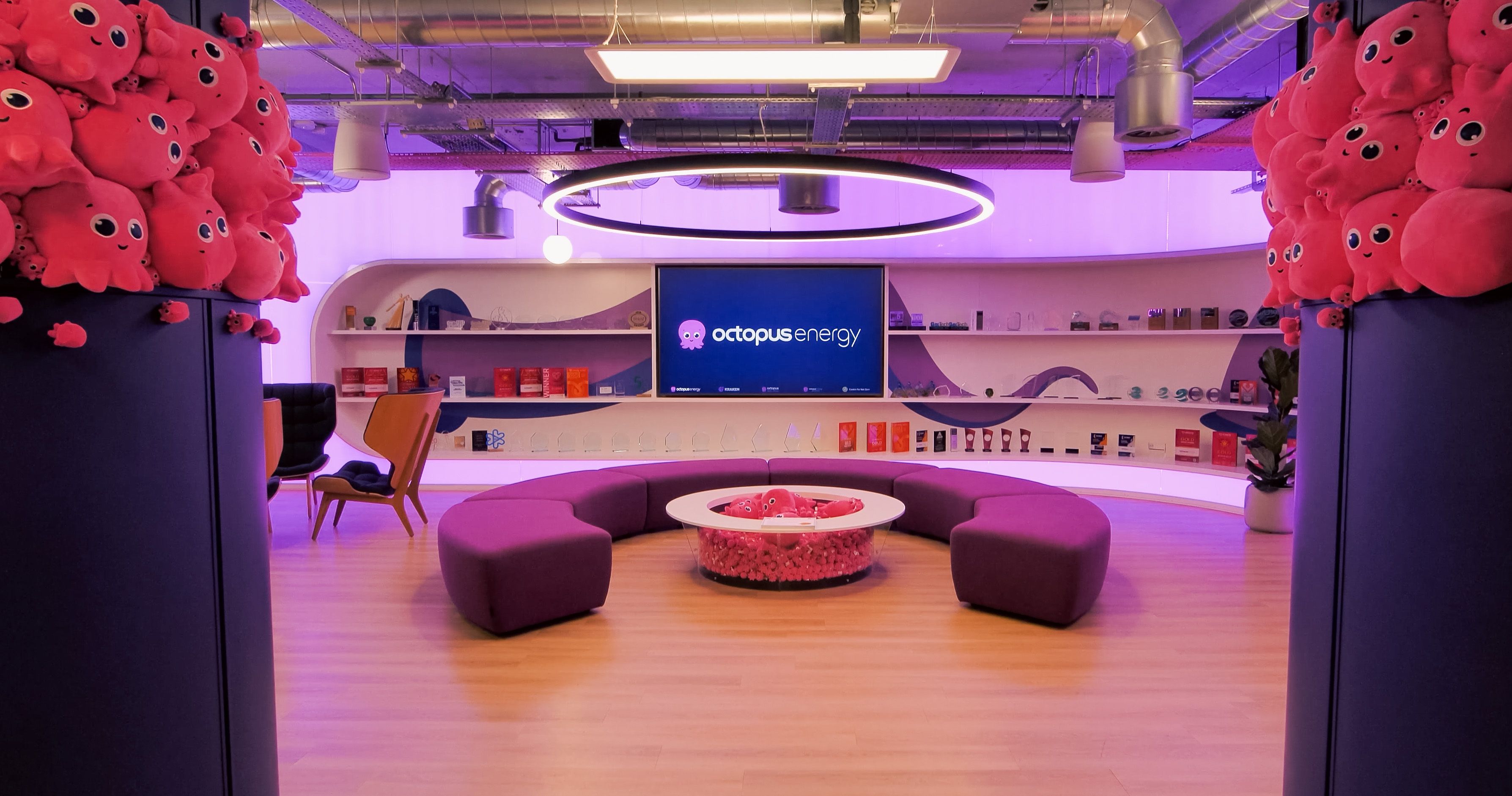 Modern lounge with an "Octopus Energy" screen, purple curved sofa, and columns covered in pink octopus plush toys.