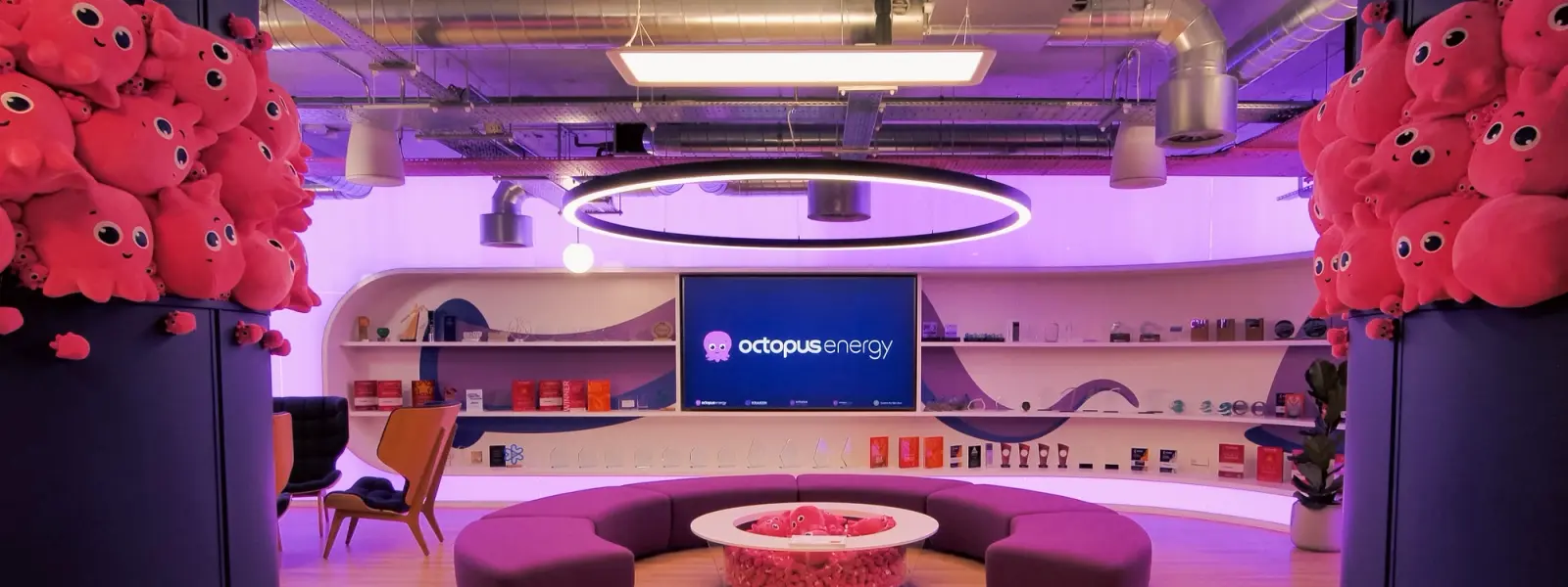 Modern lounge with an "Octopus Energy" screen, purple curved sofa, and columns covered in pink octopus plush toys.