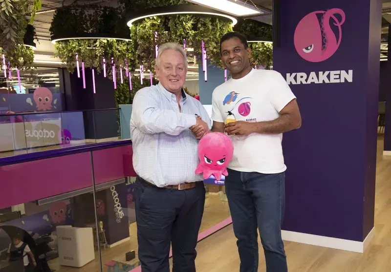 Two men, one holding a pink octopus plushie, shake hands in an office with Octopus and Kraken branding.